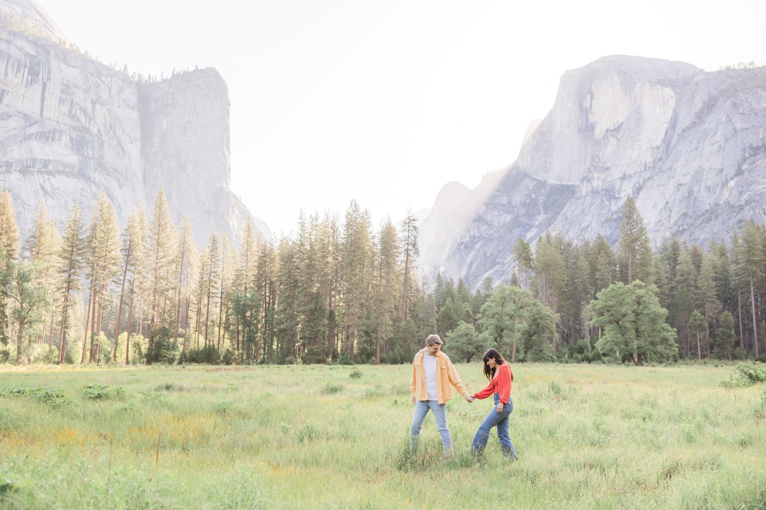 Couple holding hands walking through a green meadow in Yosemite Valley with granite cliffs and pine trees in background.