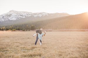 Couple dancing in golden meadow with snow-capped mountains and pine trees at sunset.