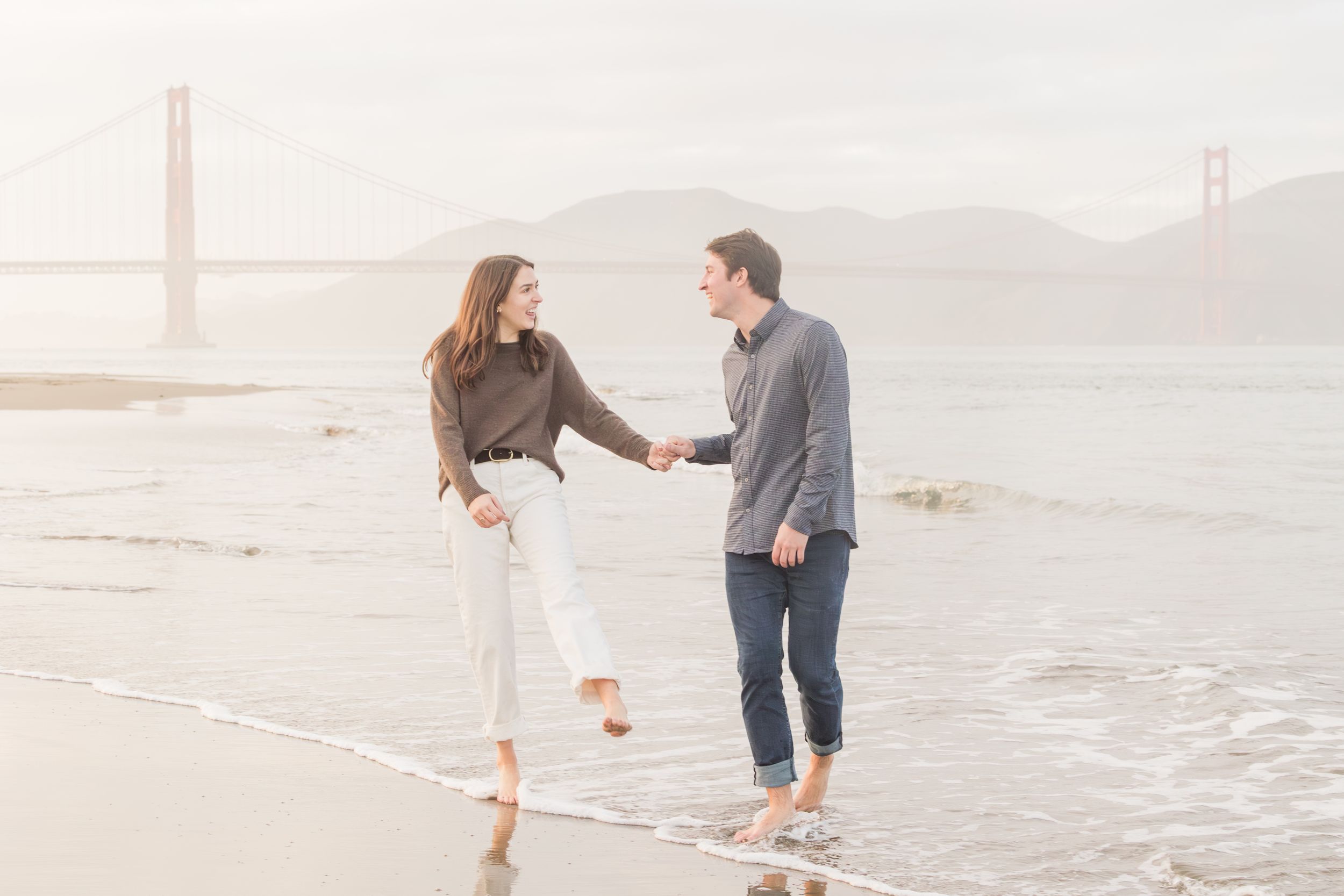 Couple holding hands and smiling on a San Francisco beach with the Golden Gate Bridge in the misty background.