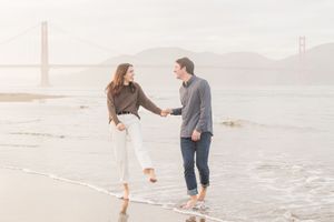Couple holding hands and smiling on a San Francisco beach with the Golden Gate Bridge in the misty background.