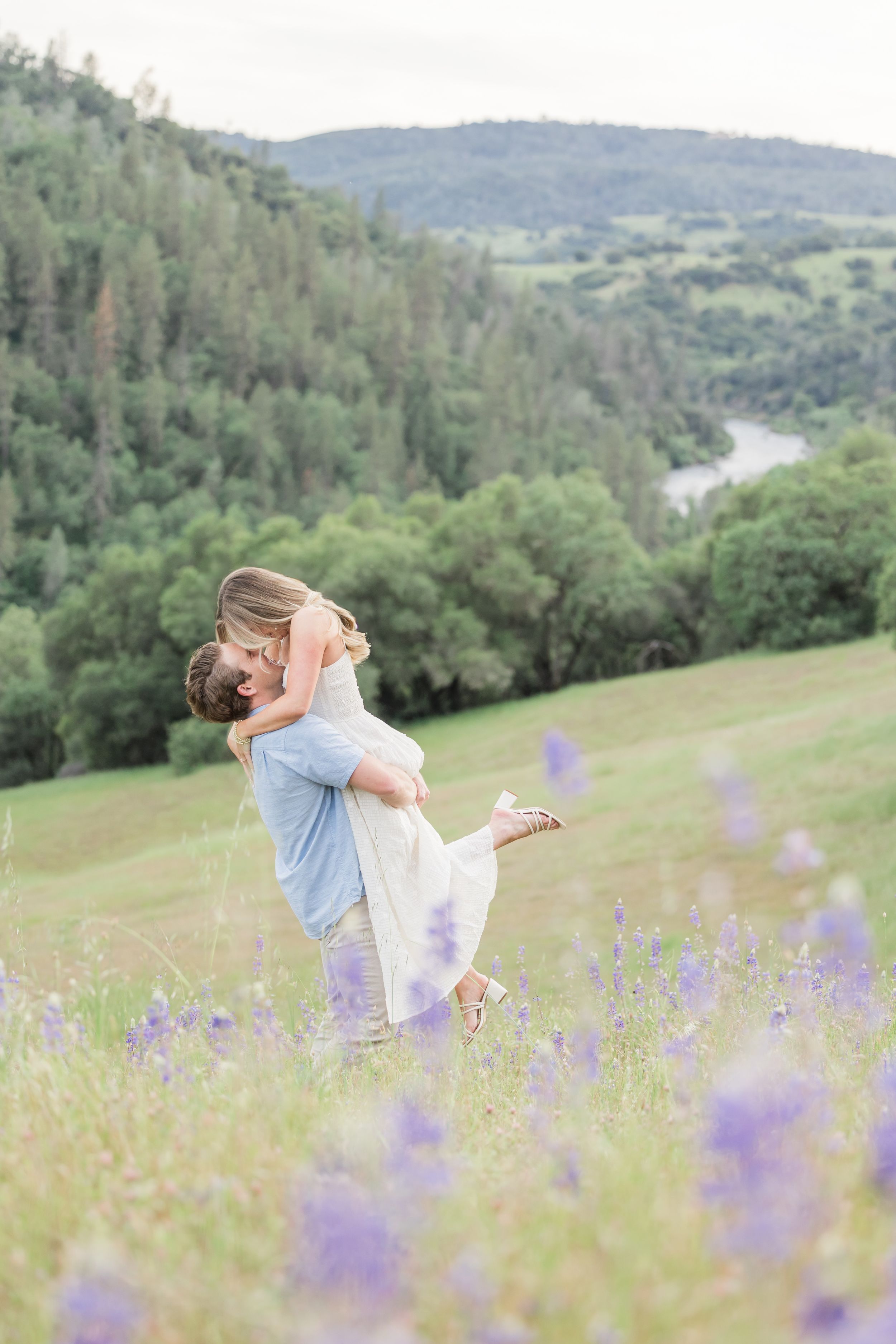 A couple shares a romantic embrace in a wildflower meadow, with lush green hills and a river in the background.