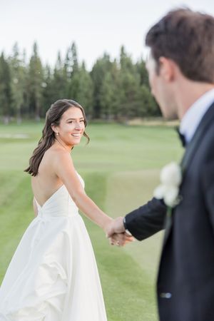 Bride in white strapless gown smiles over shoulder while holding groom's hand on lush green golf course.