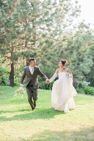 A smiling bride and groom walk hand-in-hand on green grass, surrounded by lush pine trees on their wedding day.