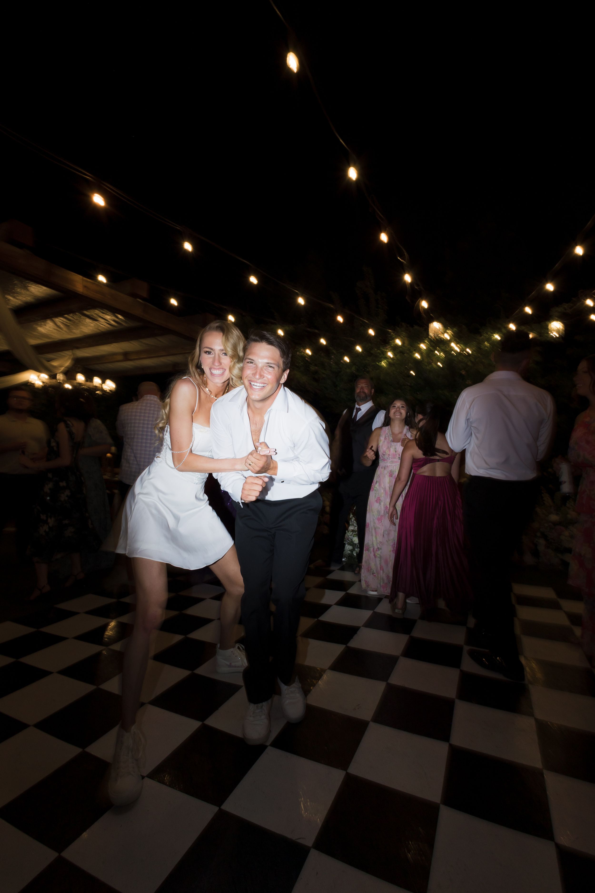 Couple dancing joyfully on checkered dance floor at outdoor evening wedding reception with string lights.
