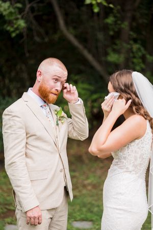Emotional bride and groom wiping tears during outdoor wedding ceremony, groom in tan suit with floral boutonniere.
