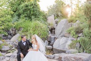 Bride and groom sit on rocks near a waterfall surrounded by lush greenery in a romantic outdoor wedding portrait.