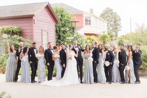 Large wedding party poses outdoors in front of a red building, bridesmaids in blue gowns and groomsmen in black tuxedos.