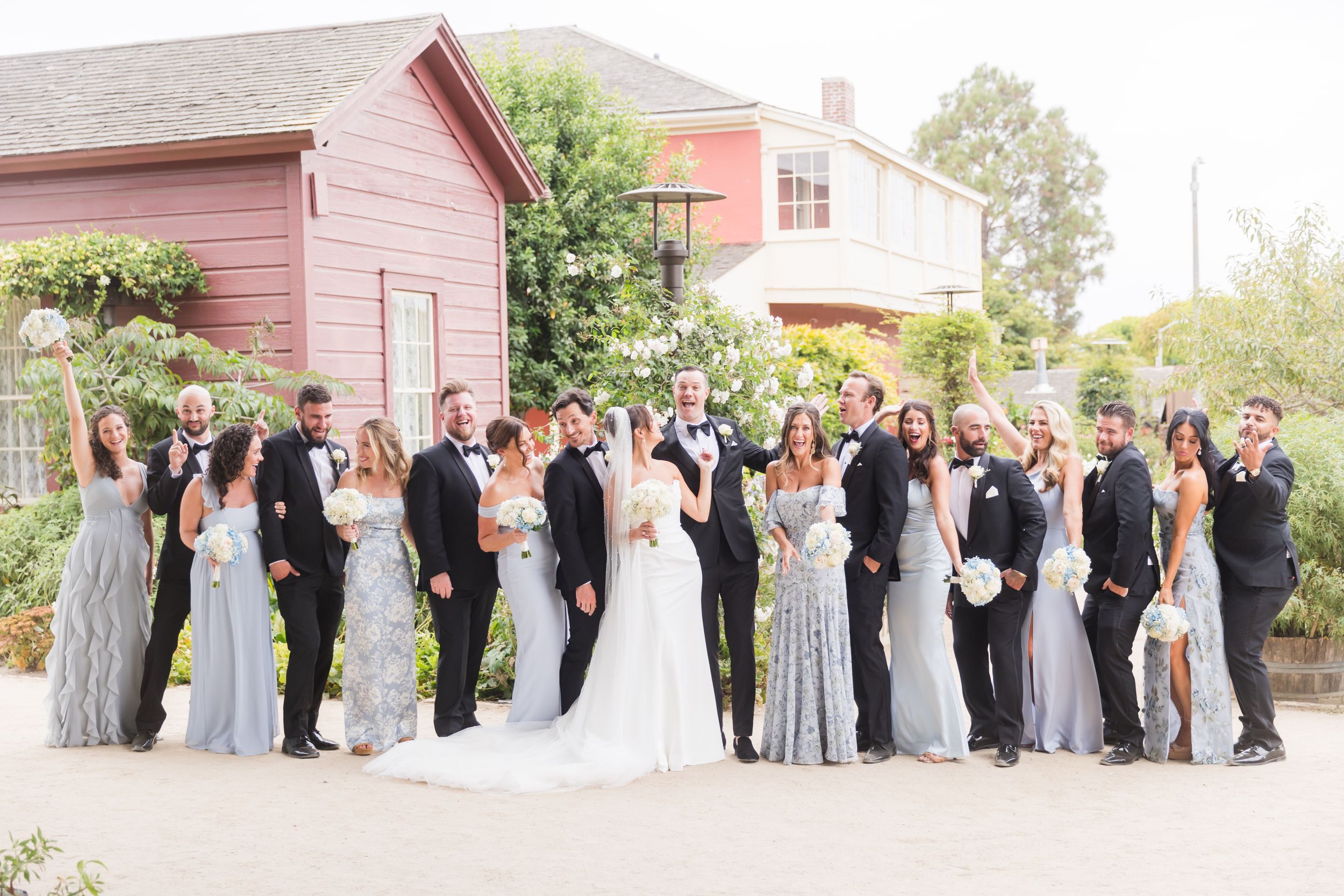 Large wedding party poses outdoors in front of a red building, bridesmaids in blue gowns and groomsmen in black tuxedos.