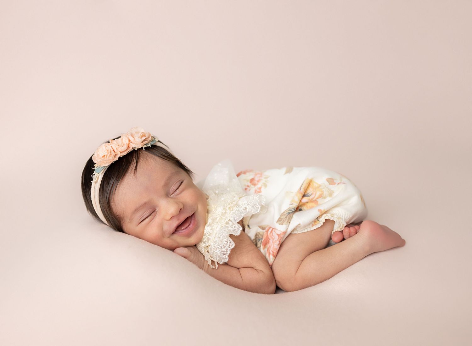 Sleeping newborn baby girl in floral romper and purple flower crown on white fluffy blanket.