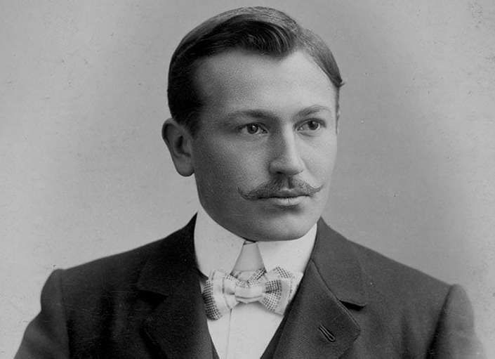 Black and white portrait of a young man with a mustache wearing a suit and bow tie, early 20th century style.