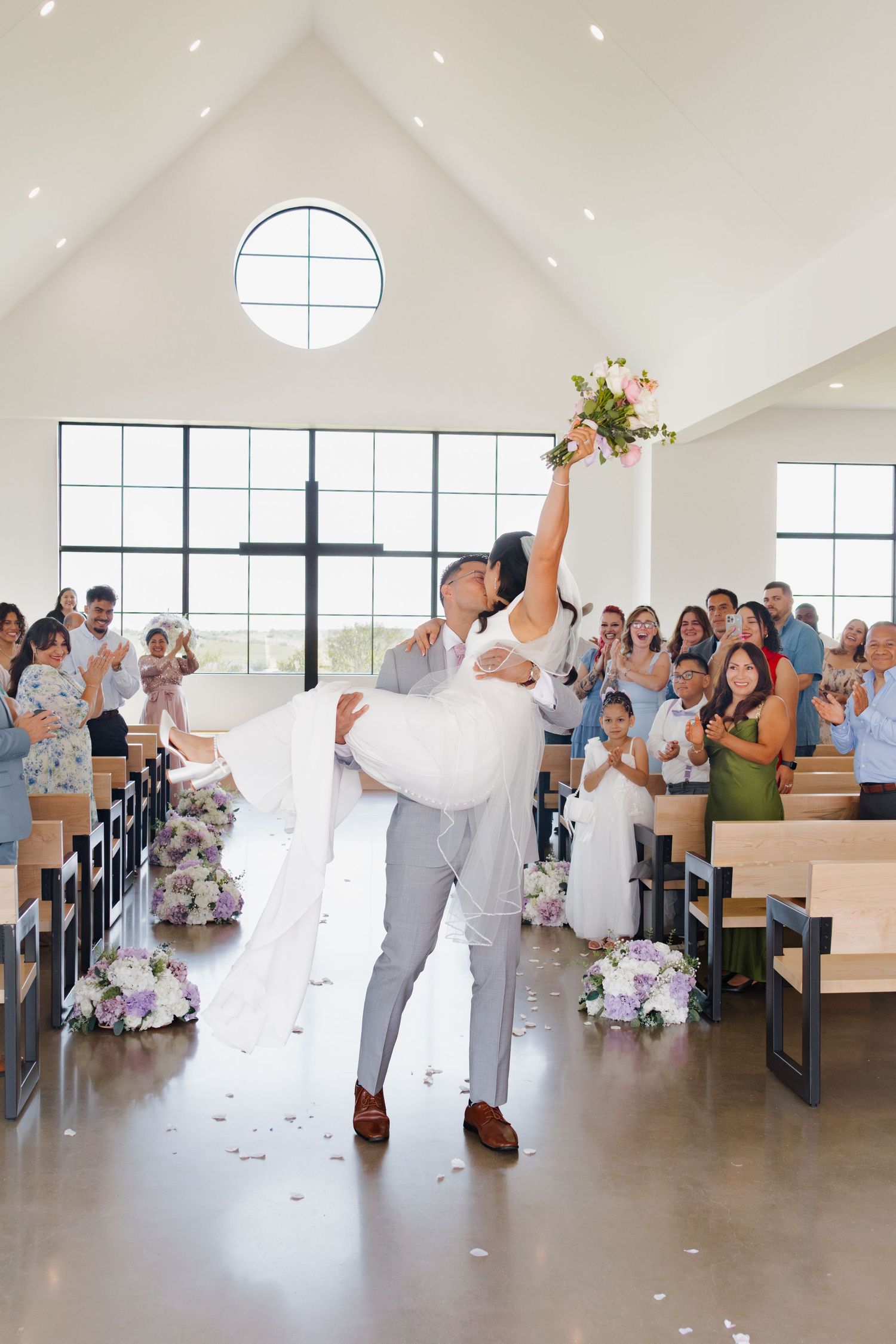 Bride feeding wedding cake to groom in gray suit at elegant reception, with tiered floral cake visible nearby.