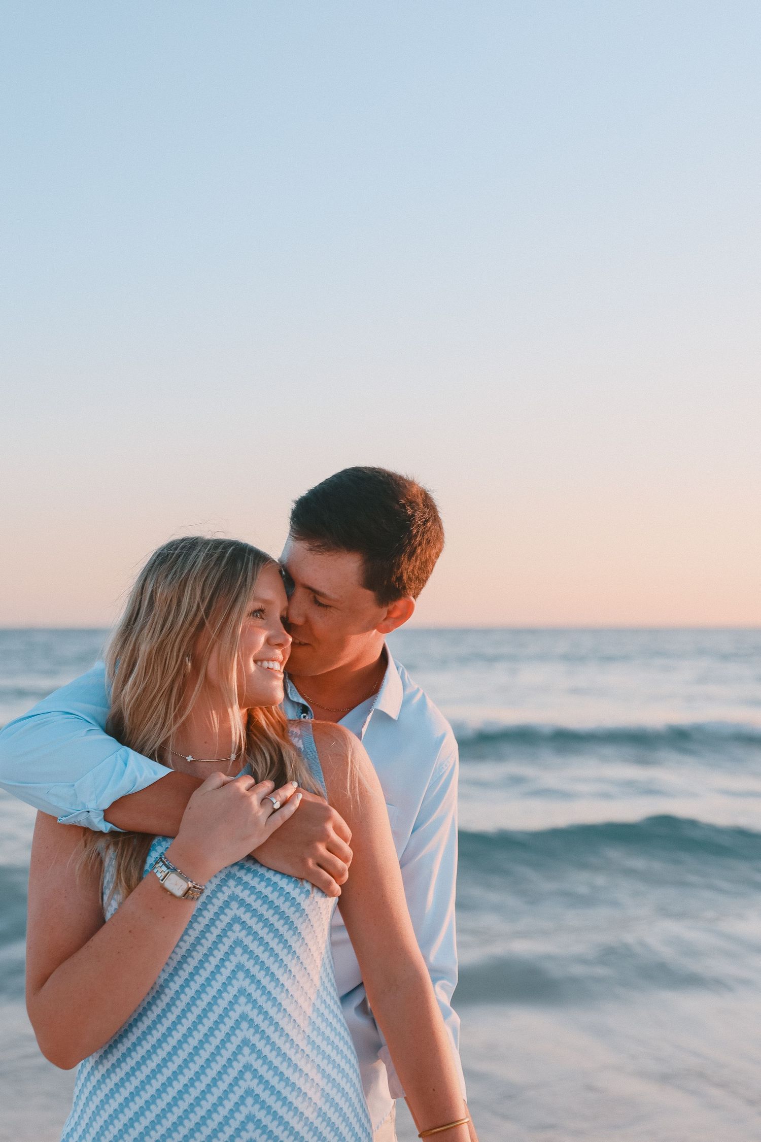 Couple embracing on beach at sunset, woman smiling in blue dress, man kissing her cheek near ocean waves.