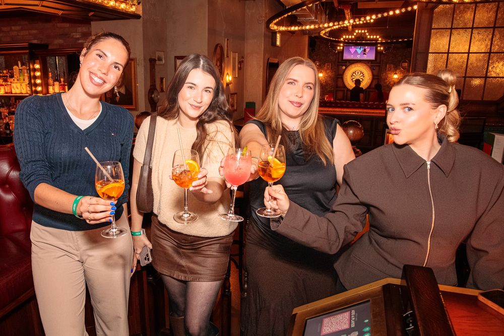 Four women smiling and toasting with colorful cocktails at a stylish bar.