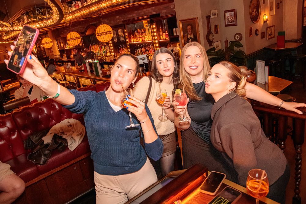 Four women taking a selfie at a bar while holding cocktails in a warmly lit pub setting.