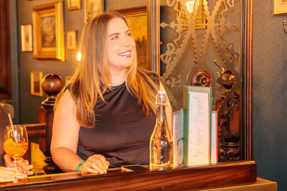 Woman in black dress smiling at a bar with a gold bottle and wine glass on the counter.