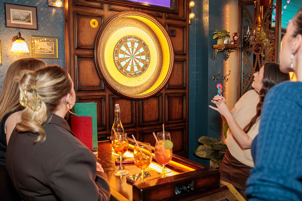 Women playing darts at a cozy bar with drinks on a wooden table and a large dartboard on the wall.