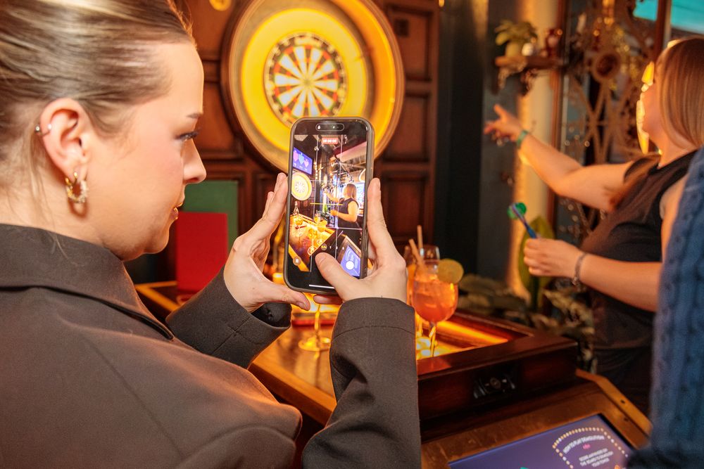 Woman photographing friends with cocktails at a bar near a dartboard.