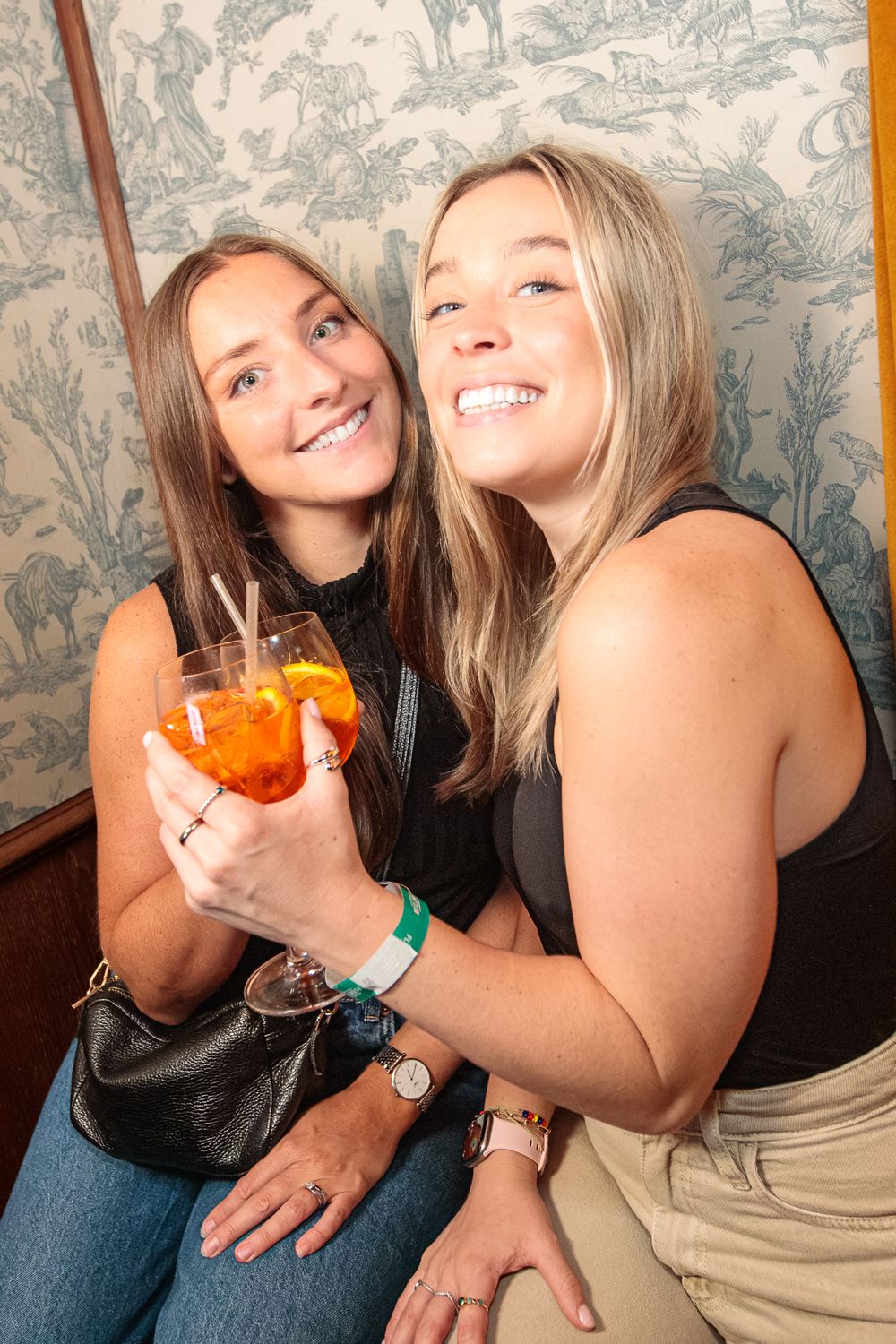 Two smiling young women posing together at a bar, one holding an Aperol Spritz cocktail with orange slice.