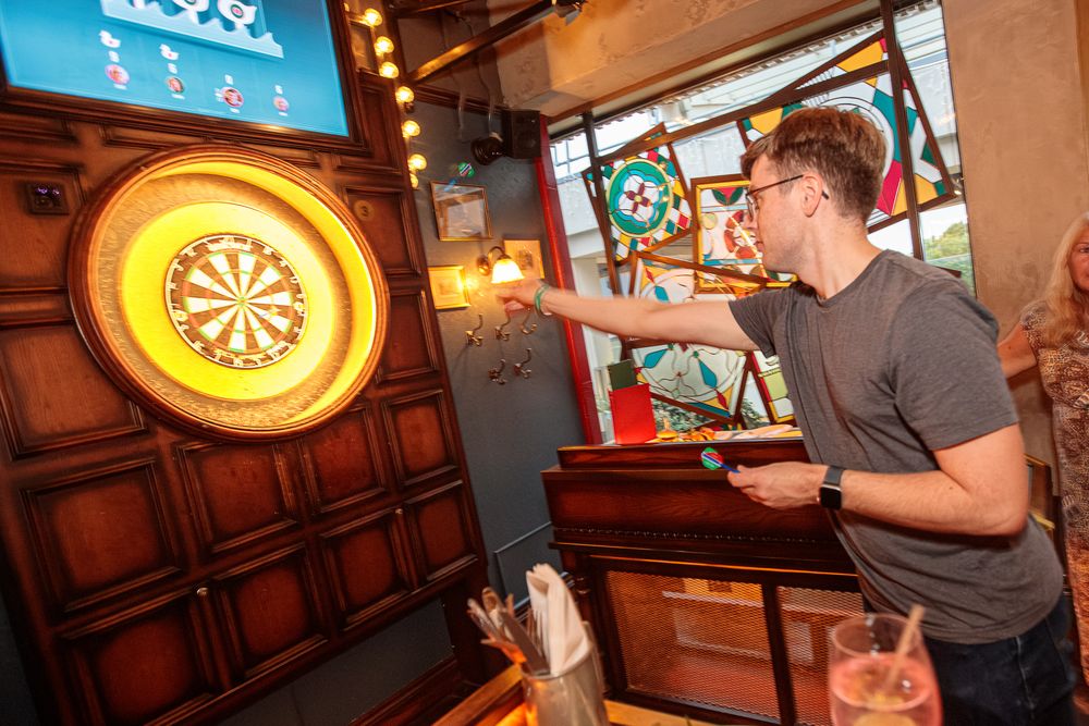 Man throwing darts at a dartboard in a pub-style bar with stained glass windows and drinks on the counter.