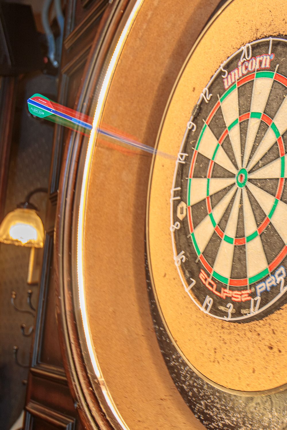 A colorful dart lands on a Unicorn Eclipse Pro dartboard mounted in an ornate wooden cabinet in a pub.