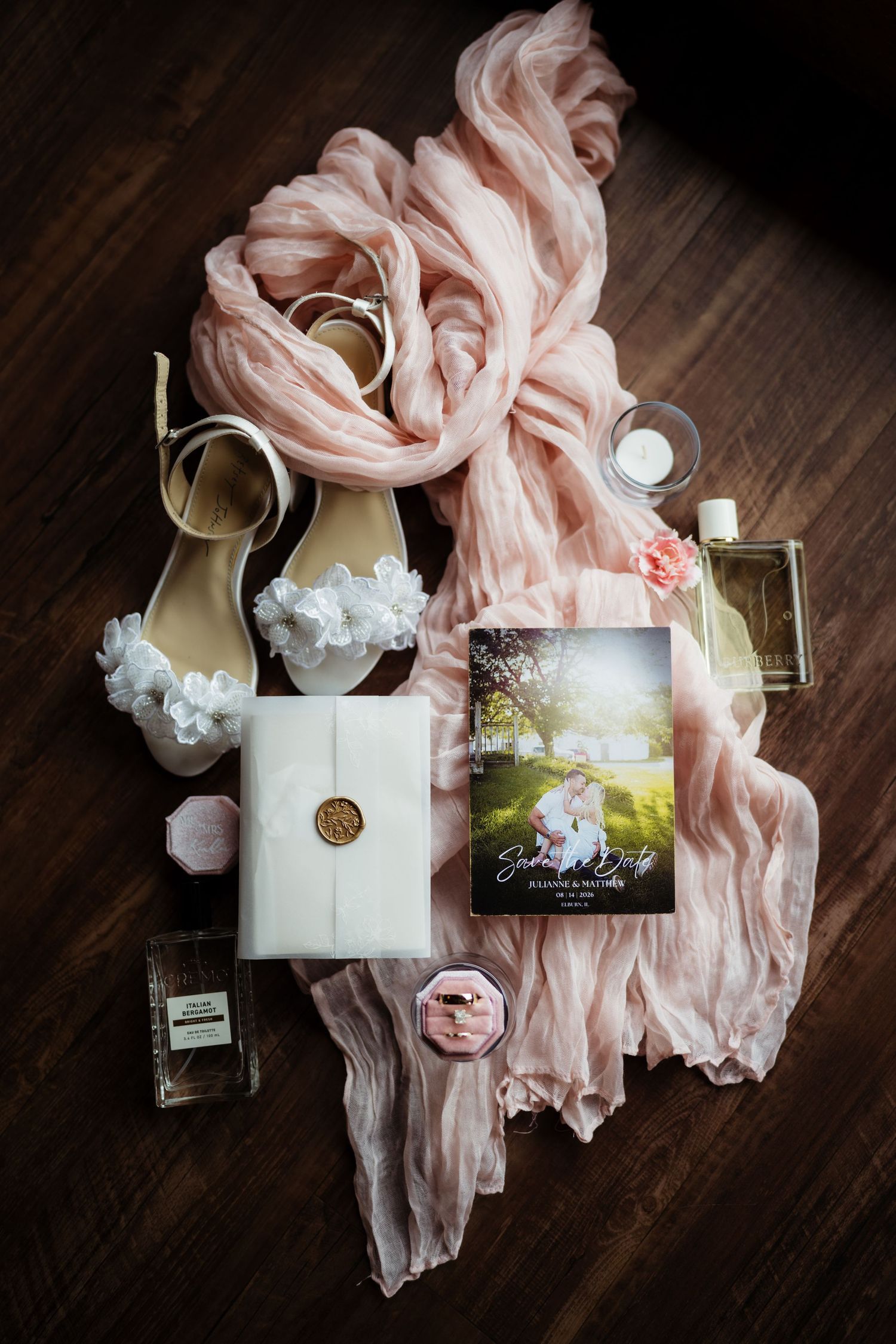 Elegant wedding flat lay with blush scarf, white heels, gold-sealed gift, perfume, and a romantic save-the-date card on dark wood.