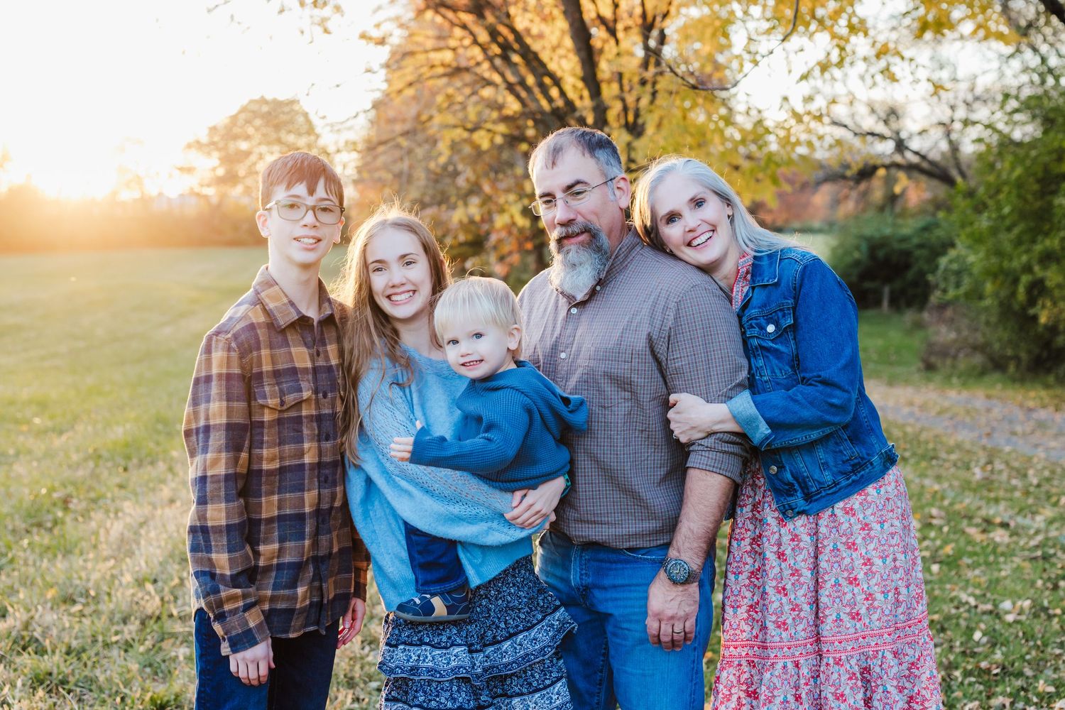 Family of five posing outdoors at golden hour with autumn trees in background.