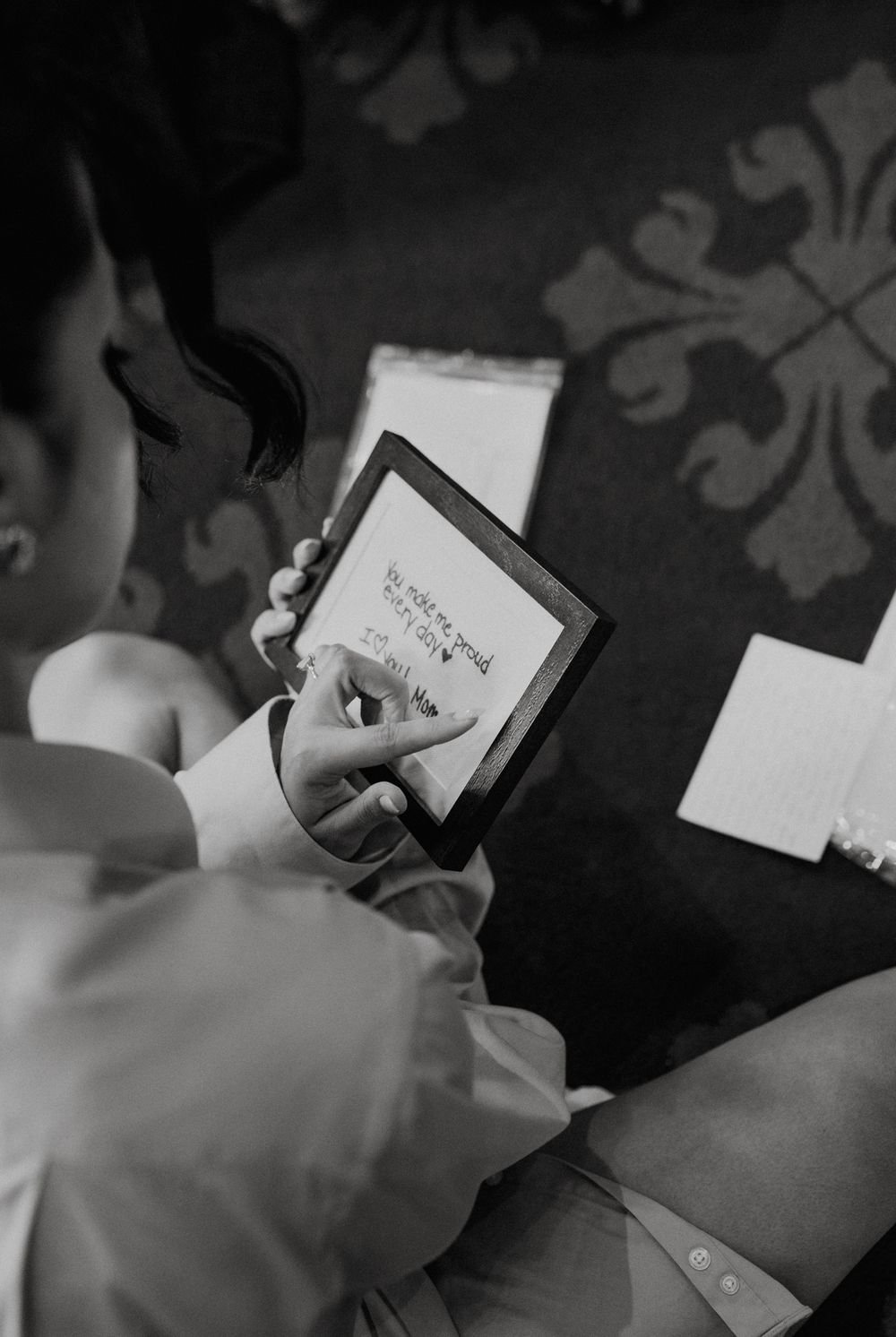 Black and white photo of a person reading a handwritten note on a small framed card during a wedding preparation.