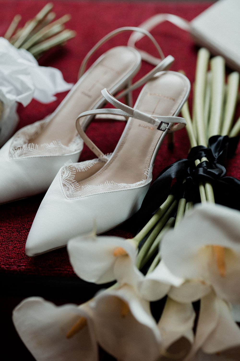 Elegant white bridal heels with ankle straps beside white calla lilies on a red surface.