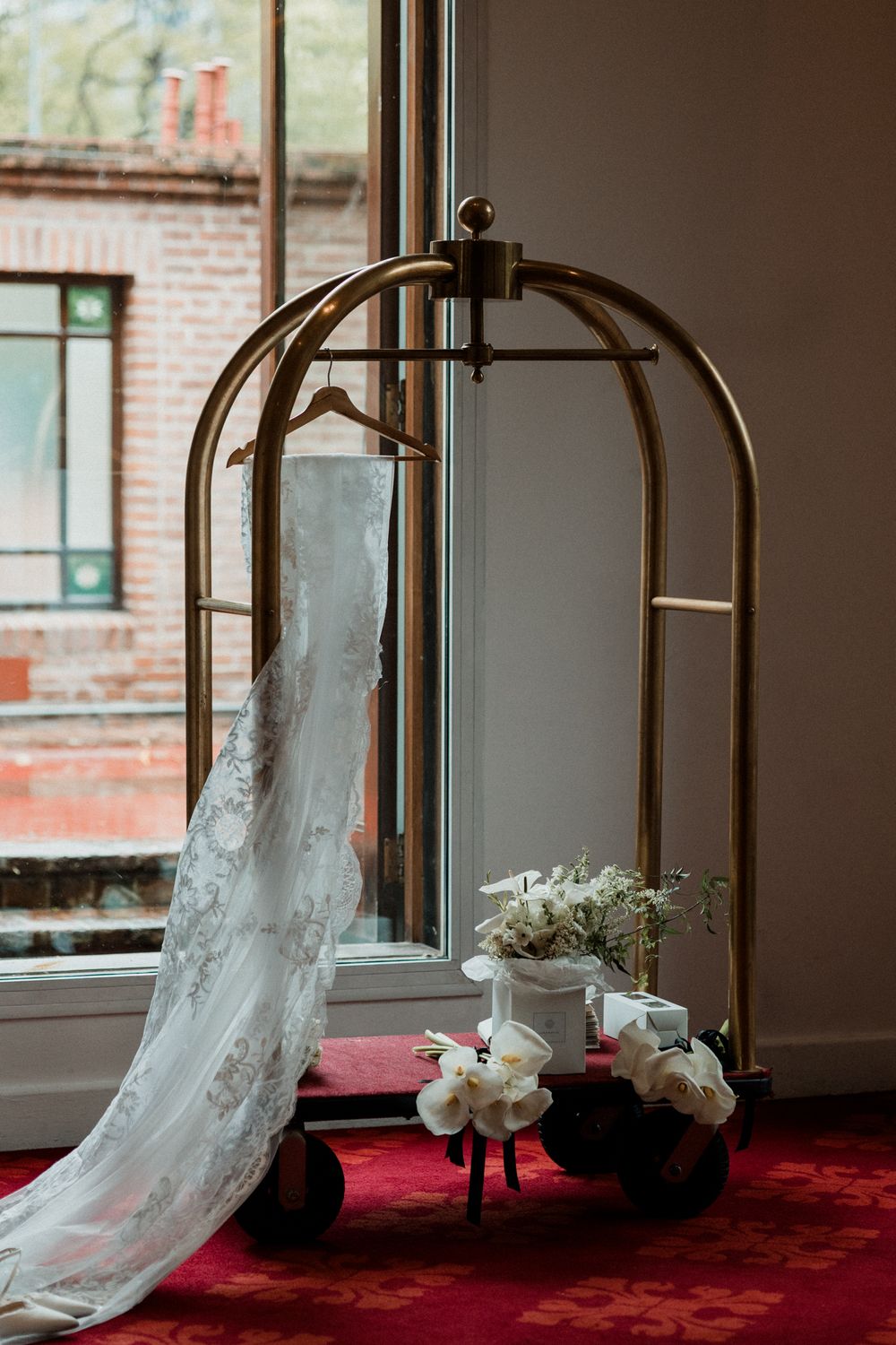 A elegant wedding dress hanging on a gold luggage cart beside white floral arrangements near a window.