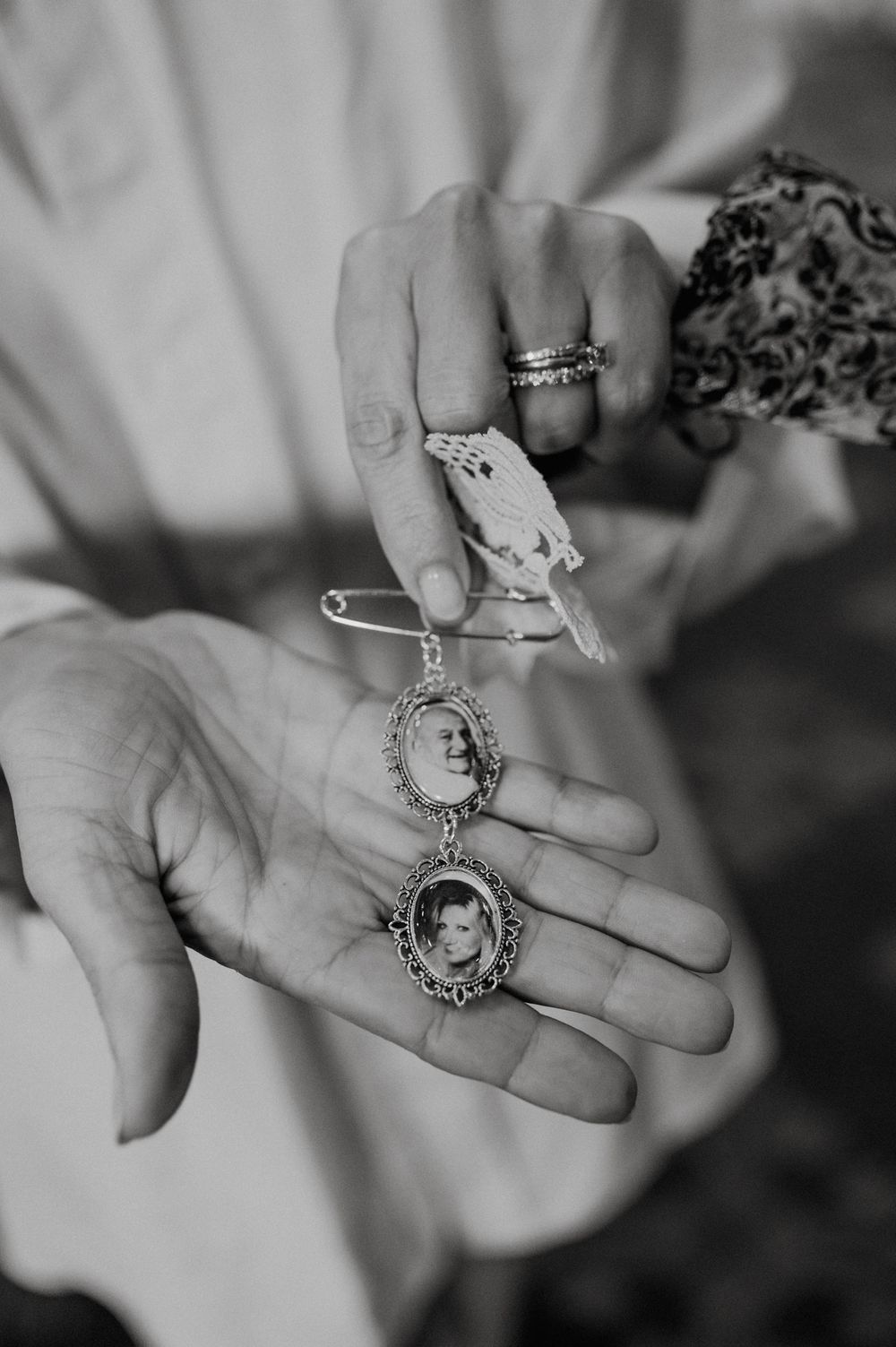 Hands holding two oval locket charms with portrait photos on a decorative pin, black and white photography.