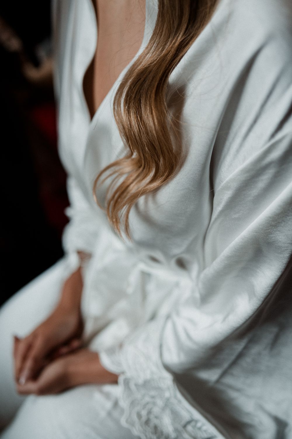 A bride in a white robe sits with hands folded, her long wavy blonde hair cascading over her shoulder.