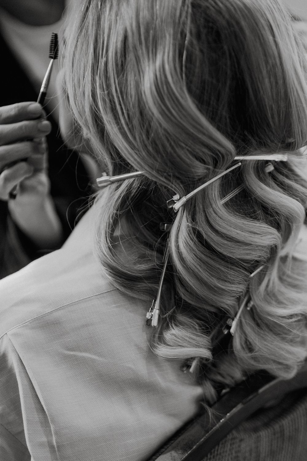 Black and white photo of a woman's curly blonde hair being styled with clips and a brush by a hairstylist.