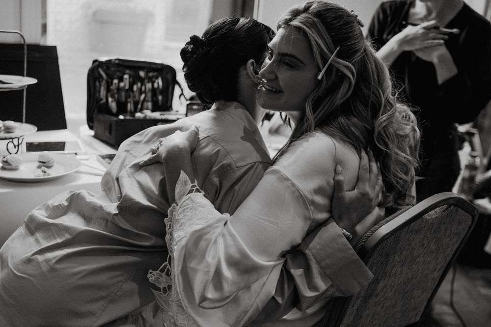 Two women in silk robes share a tender embrace while getting ready for a wedding in black and white photo.