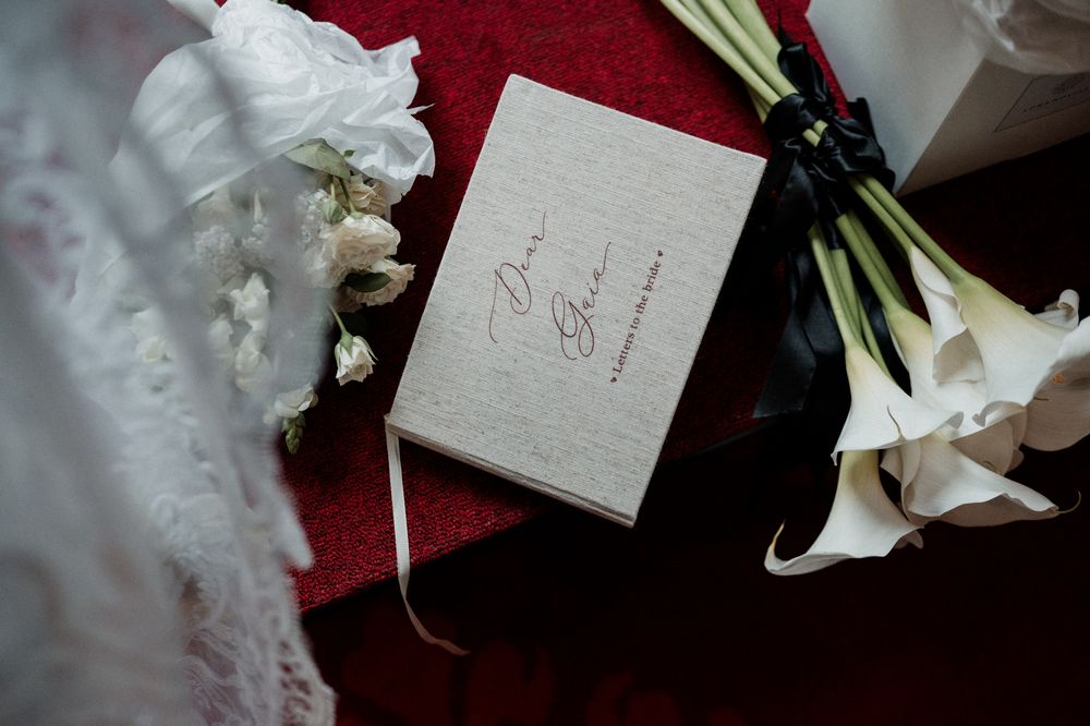 Elegant wedding flat lay with a linen vow book, white calla lily bouquet, and bridal lace on red velvet.