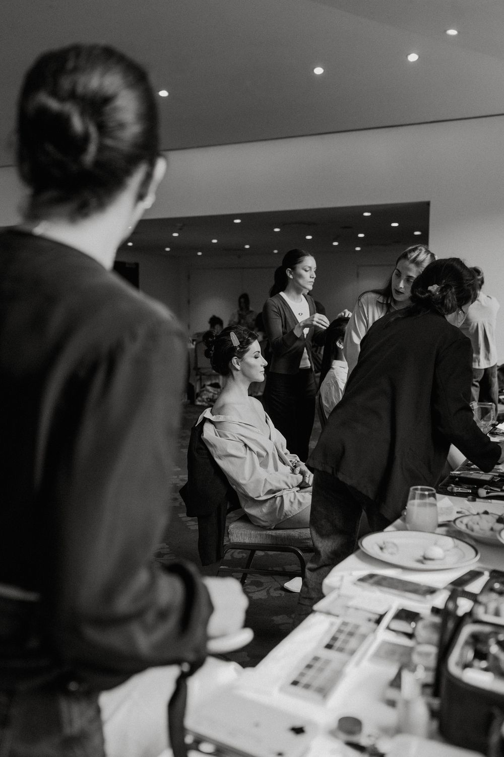 Black and white photo of a bride getting hair and makeup done by stylists in a bright modern bridal suite.