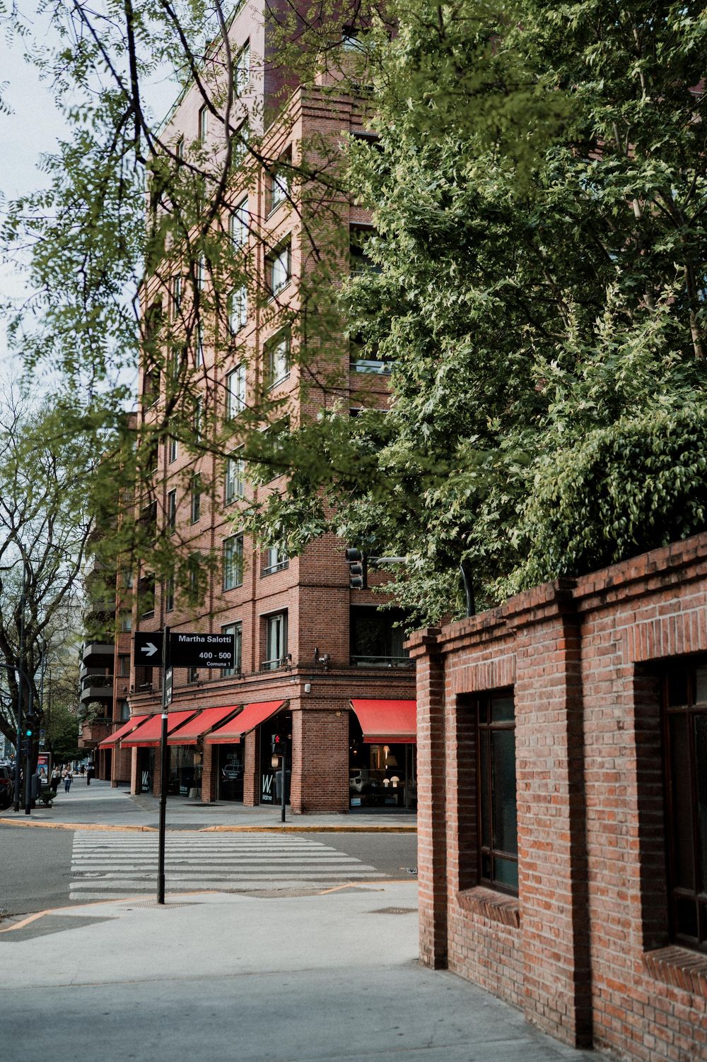 Historic brick building with red awnings and lush green trees lining an urban sidewalk on a sunny day.