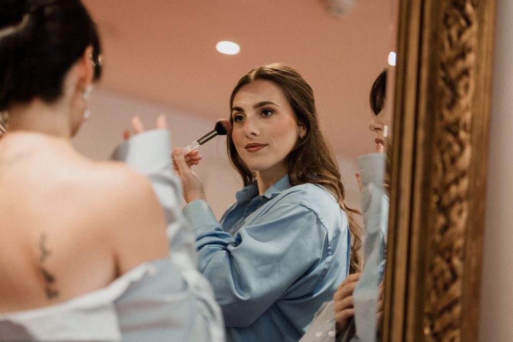 Woman applying blush with makeup brush while looking in ornate gold mirror during bridal getting-ready session.