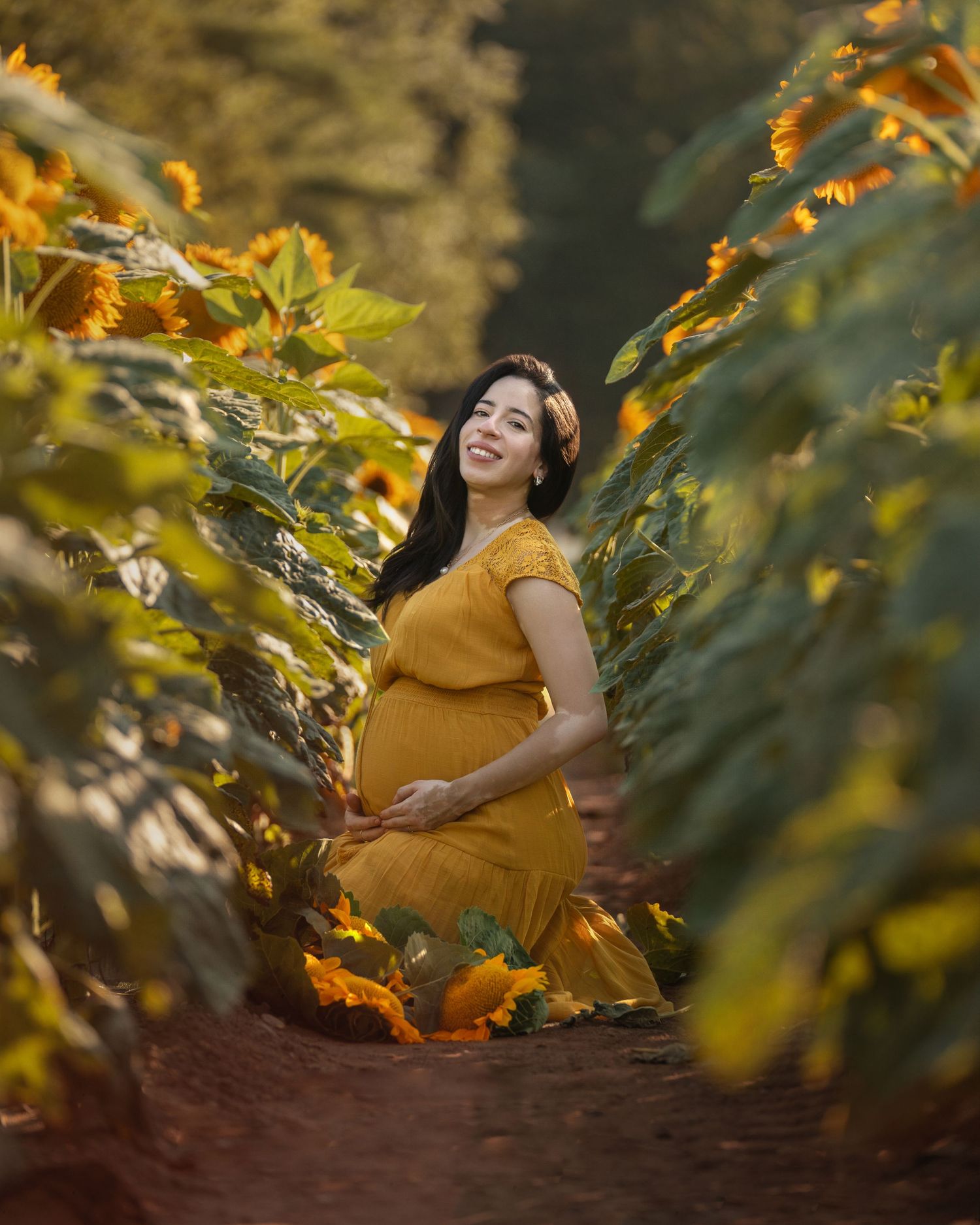 Pregnant woman in yellow dress smiling among sunflowers in a field, cradling her baby bump.