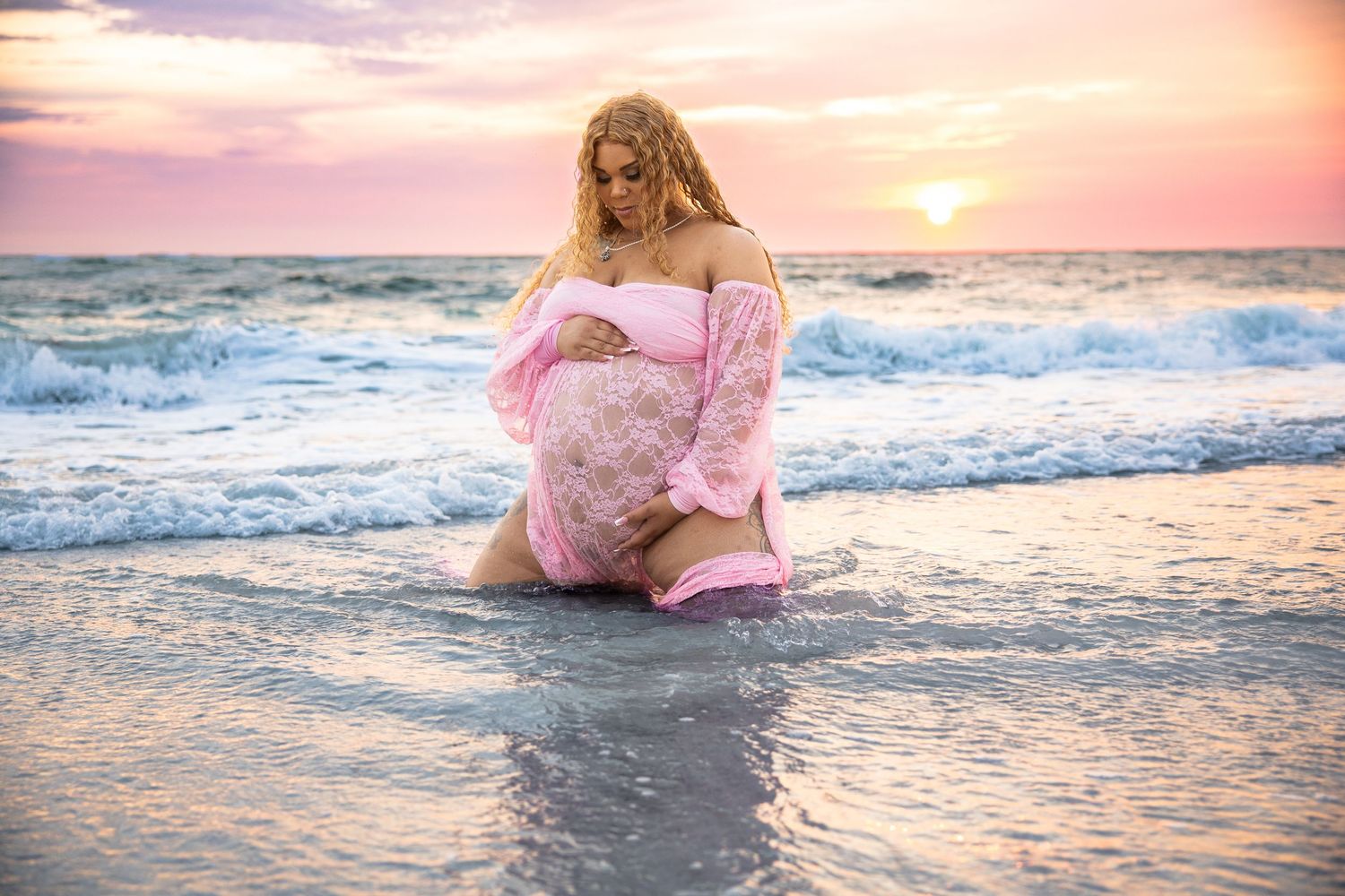 Pregnant woman in pink lace gown kneeling on beach at sunset, cradling baby bump with ocean waves.