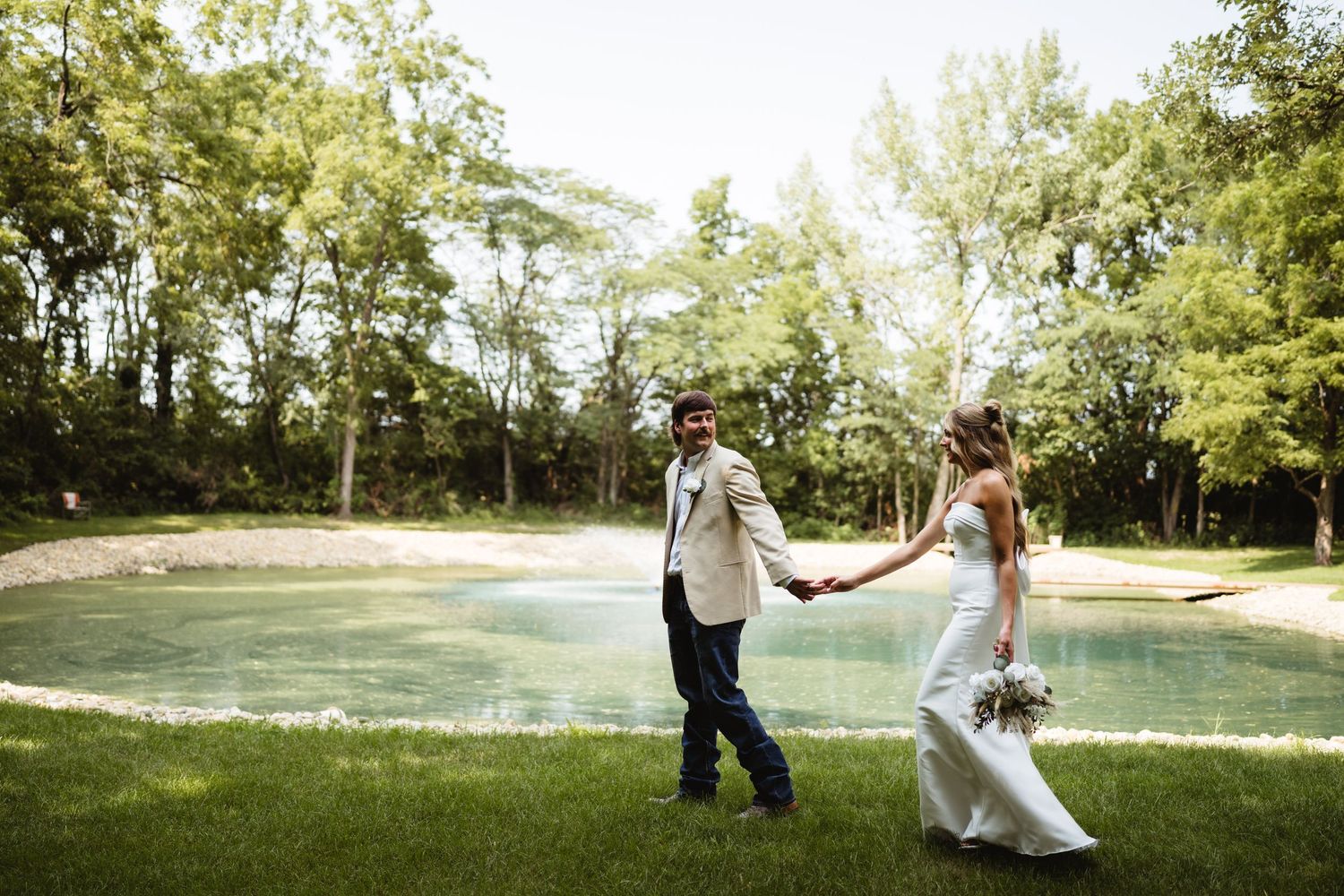 Bride and groom hold hands walking by a serene pond surrounded by lush green trees on their wedding day.
