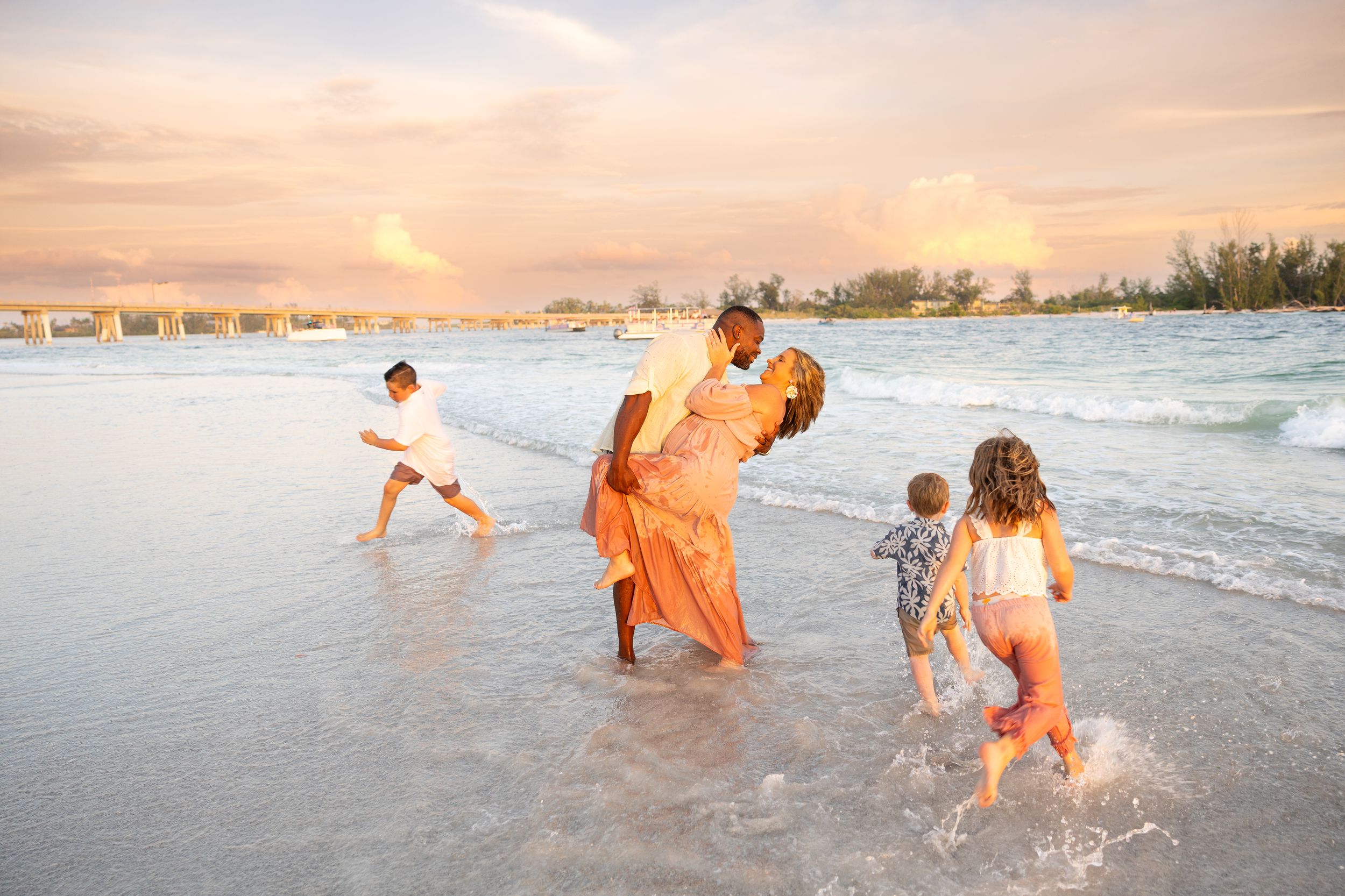 Family playing in ocean waves at sunset on a tropical beach, with a colorful pink and orange sky.