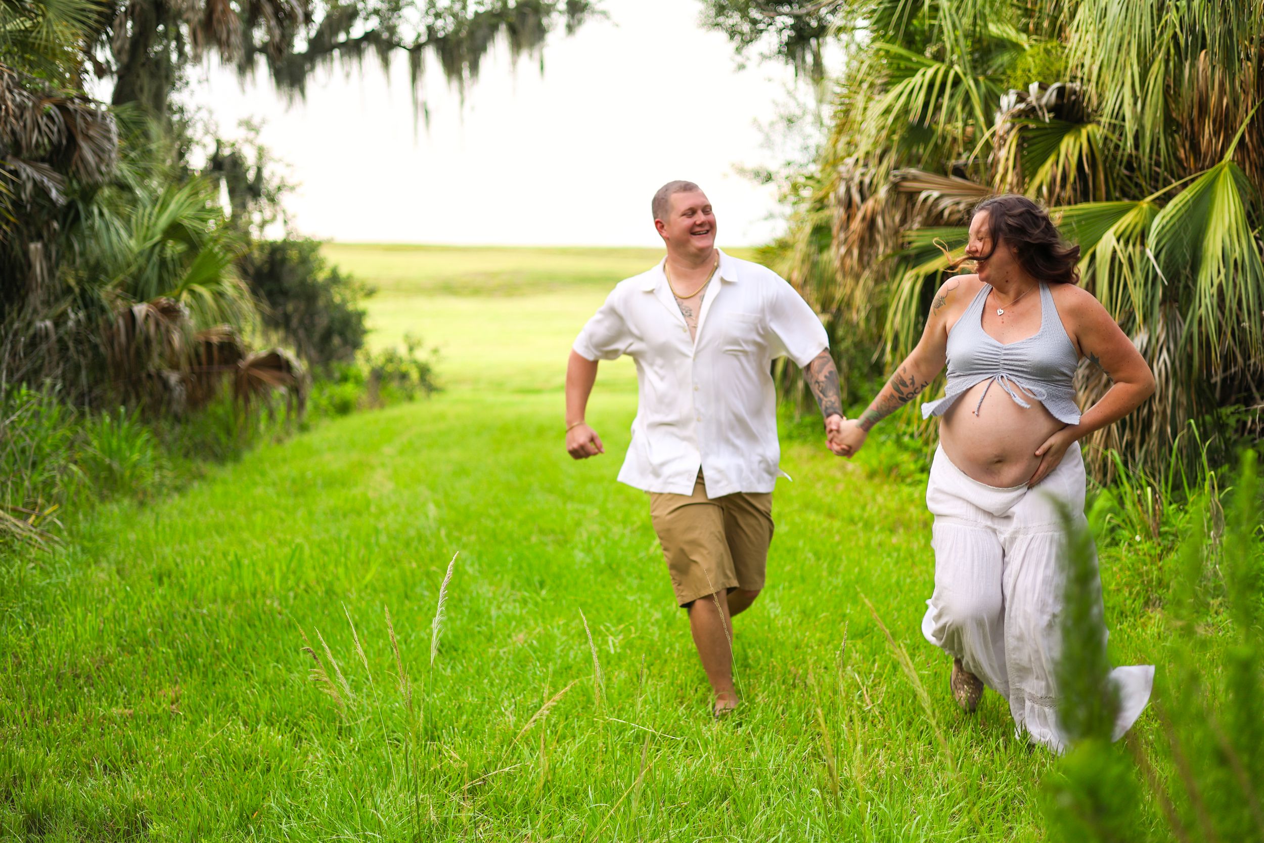 Pregnant couple holding hands running through lush green field surrounded by tropical trees during maternity photoshoot.