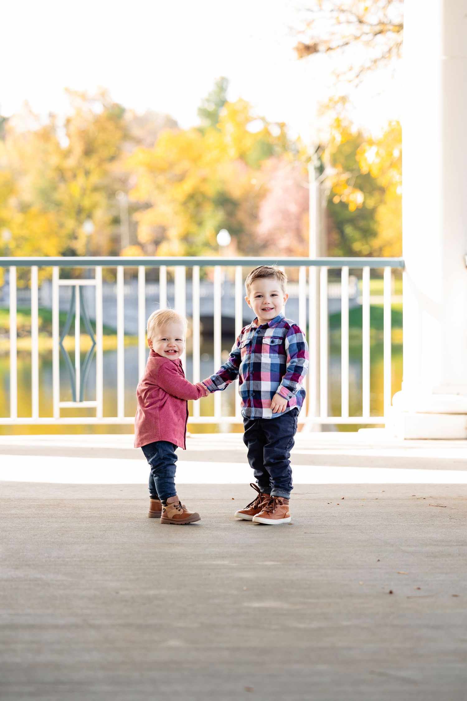 Two young children holding hands outdoors in fall, wearing plaid and pink jackets with jeans and brown boots.