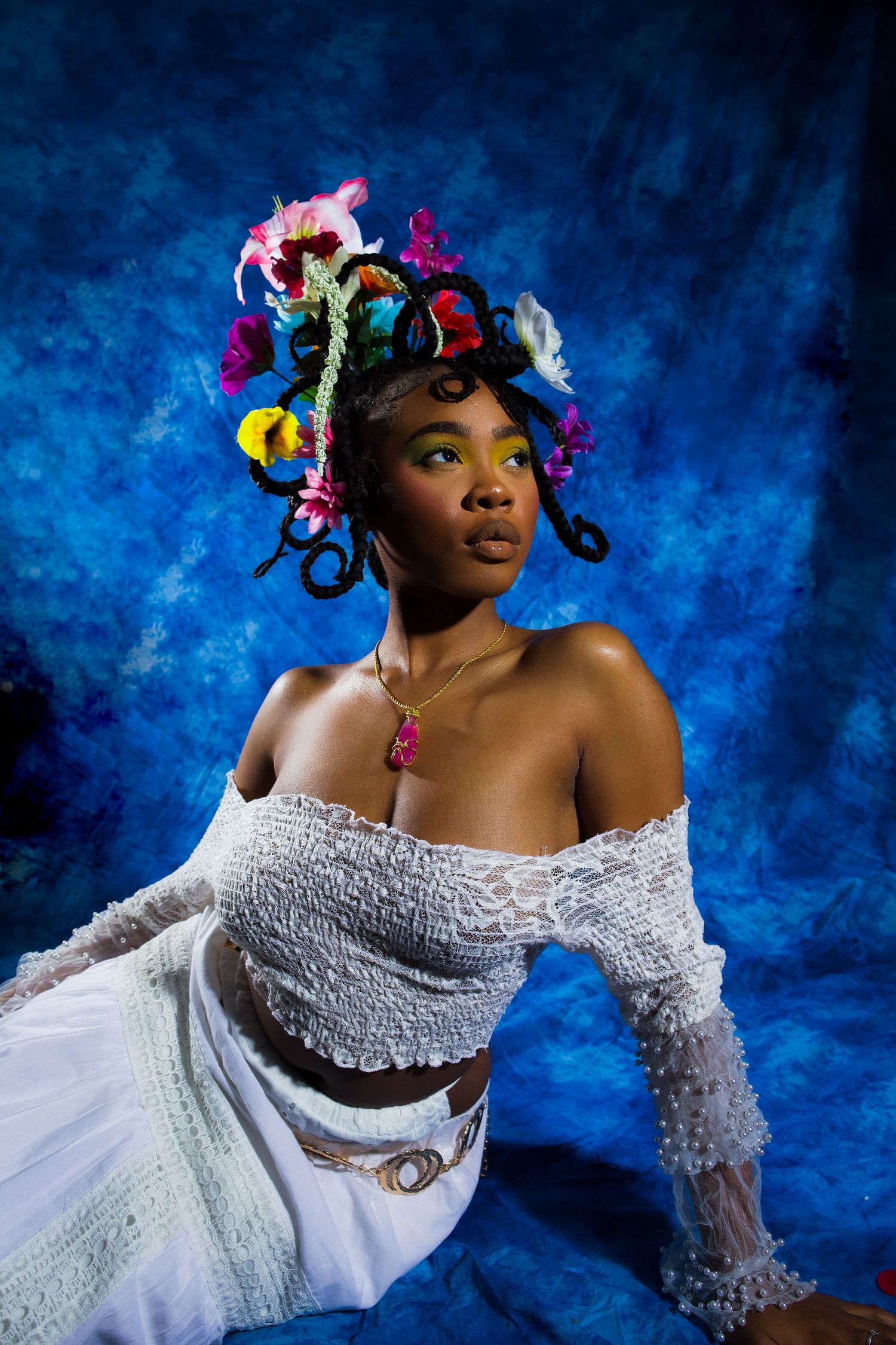 Woman in white off-shoulder top with colorful floral headpiece poses against blue textured background.