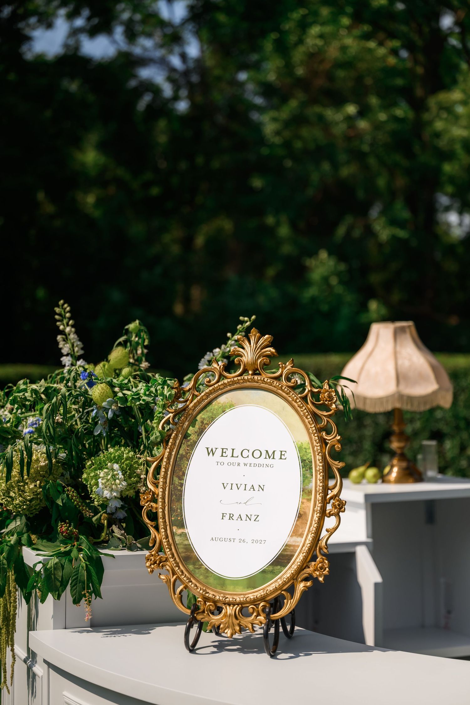 Gold ornate framed wedding welcome sign for Vivian and Franz, displayed outdoors with lush greenery and floral arrangements.