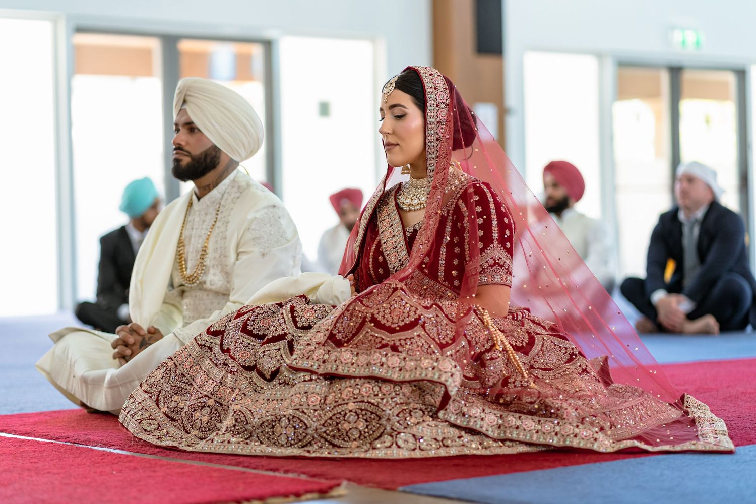 Sikh bride and groom in traditional attire seated during wedding ceremony, bride in red lehenga with dupatta.