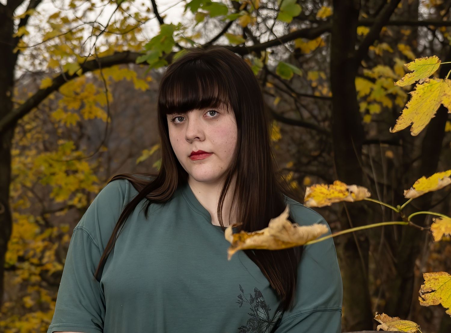 Young woman with dark hair and bangs in green top holding autumn leaves in a forest setting.