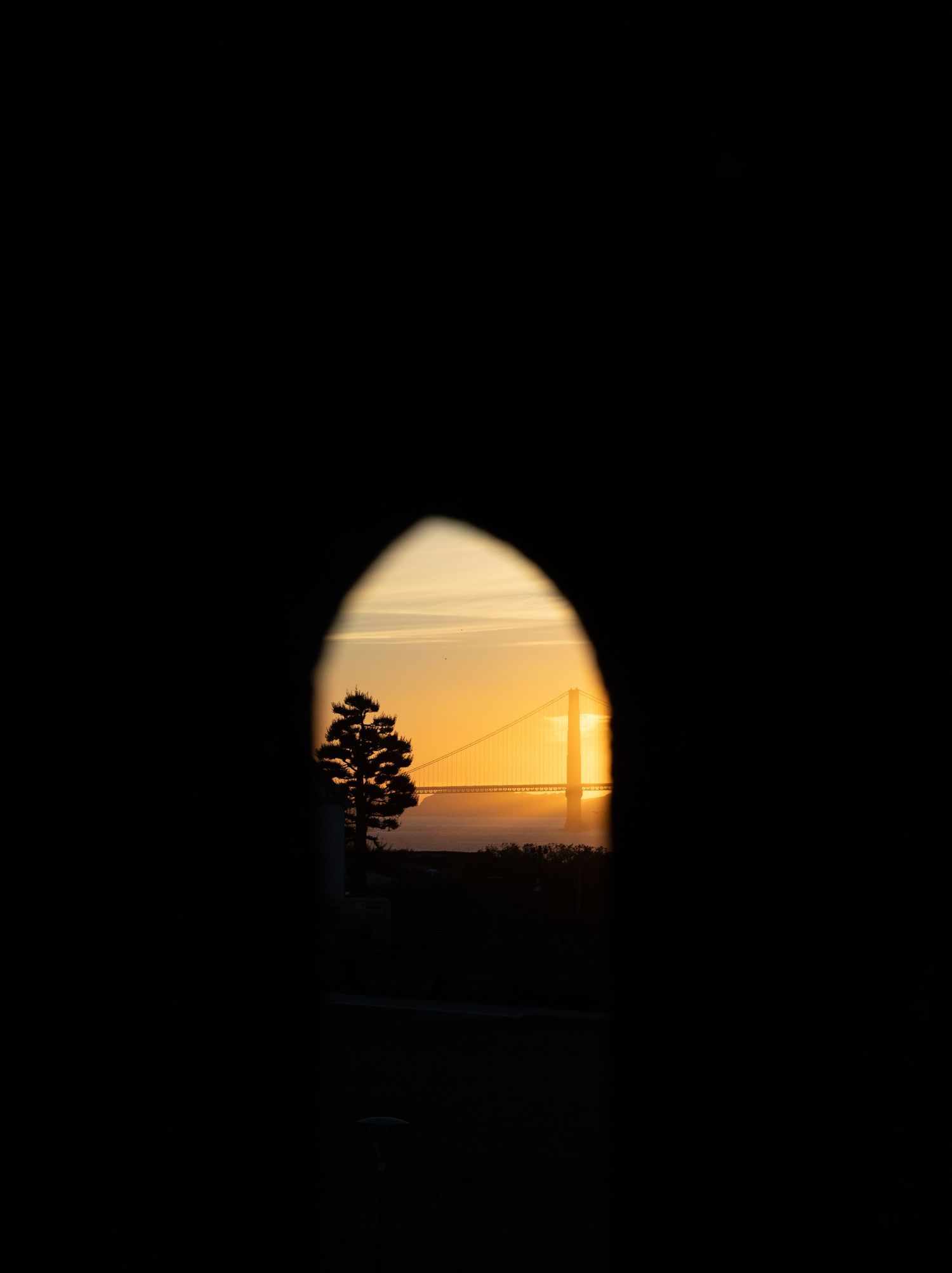 Silhouette of Golden Gate Bridge at sunset viewed through a dark arched opening with a tree silhouette.