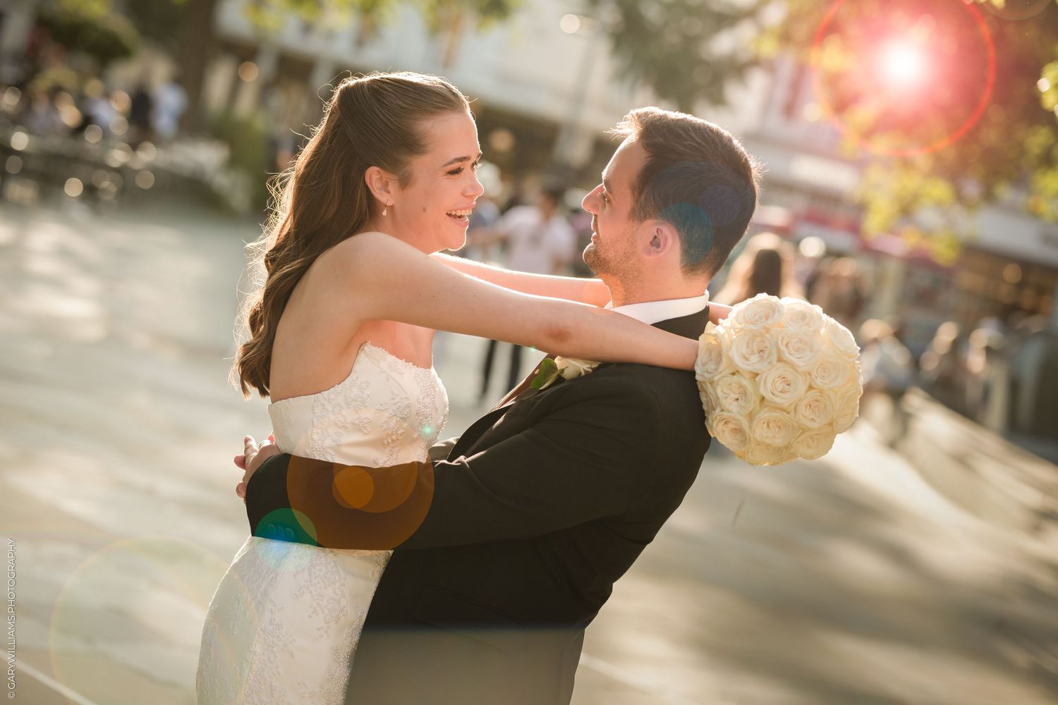 A bride and groom share a joyful embrace outdoors, with the bride holding a white bouquet in warm golden sunlight.