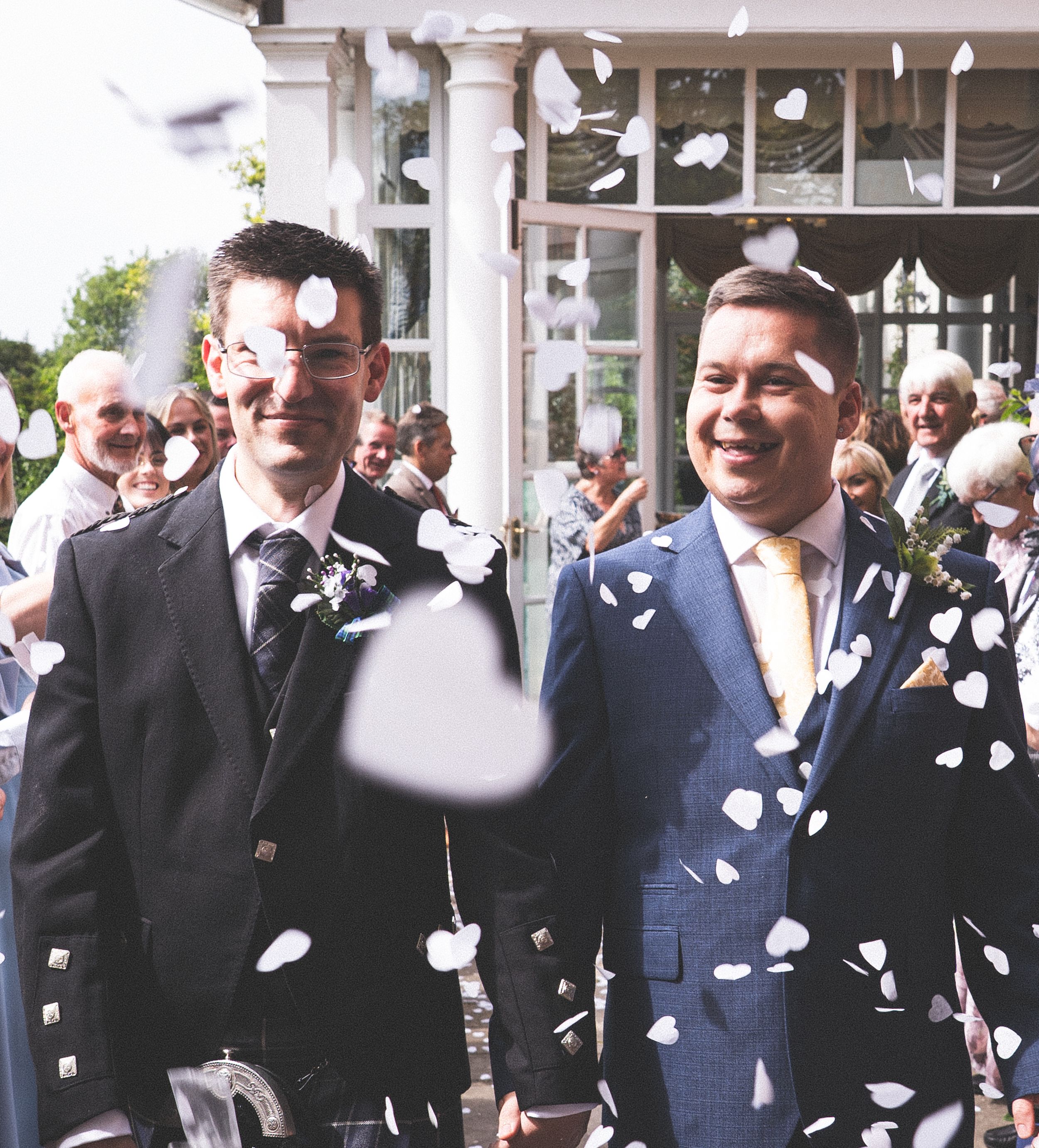 Two grooms smiling as wedding confetti falls during their outdoor civil ceremony celebration.